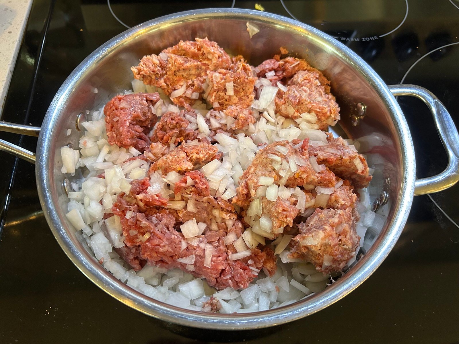 Uncooked ground beef, ground sausage , and diced onions in a steel pan in the stove.